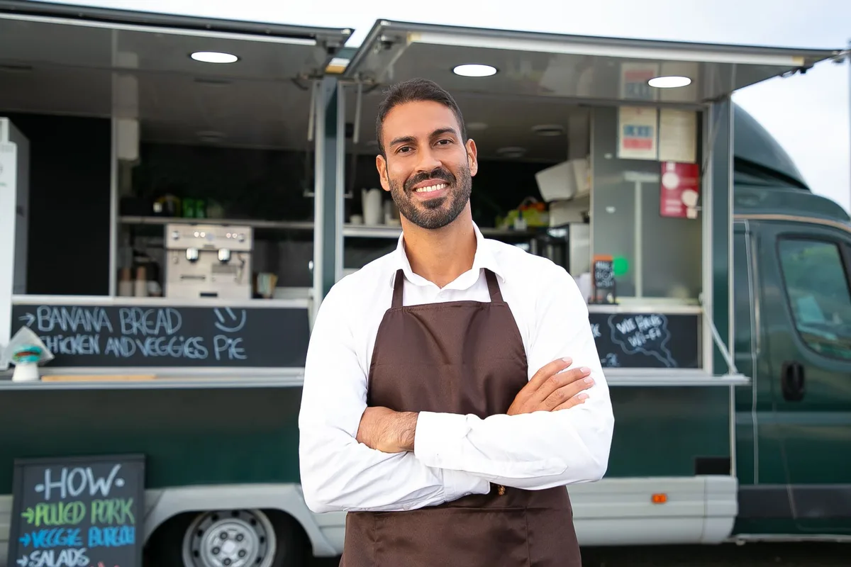 El dueño de un food truck, en camisa blanca y mandil marrón, posa sonriendo con los brazos cruzados frente a su truck verde oscuro con la ventanilla abierta. En las pizarras a su alrededor se lee BANANA BREAD, CHICKEN AND VEGGIES PIE, WE HAVE WI-FI, y un menú con PULLED PORK, VEGGIE BURGER y SALADS.